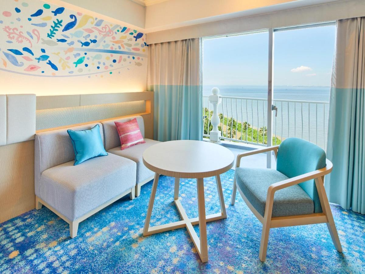 Hotel Grand Nikko Tokyo Bay Maihama Urayasu Exterior foto The photo shows a modern and stylish interior of a room with a view. There is a cozy seating area featuring a light-colored sofa with decorative cushions in various shades of blue and pink. A round table occupies the center of the space, surrounded b