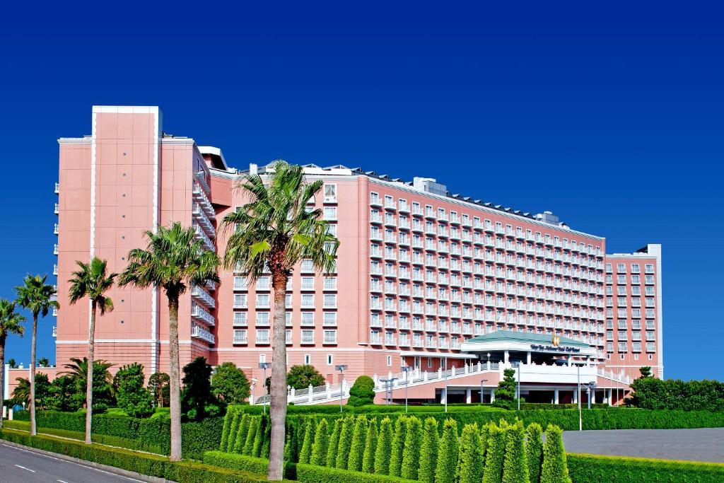 Hotel Grand Nikko Tokyo Bay Maihama Urayasu Exterior foto The image shows a large, multi-story hotel building featuring a light pink facade. The architecture appears modern, with numerous windows arranged in a grid pattern. In the foreground, there are neatly trimmed shrubs and palm trees, adding a tropical
